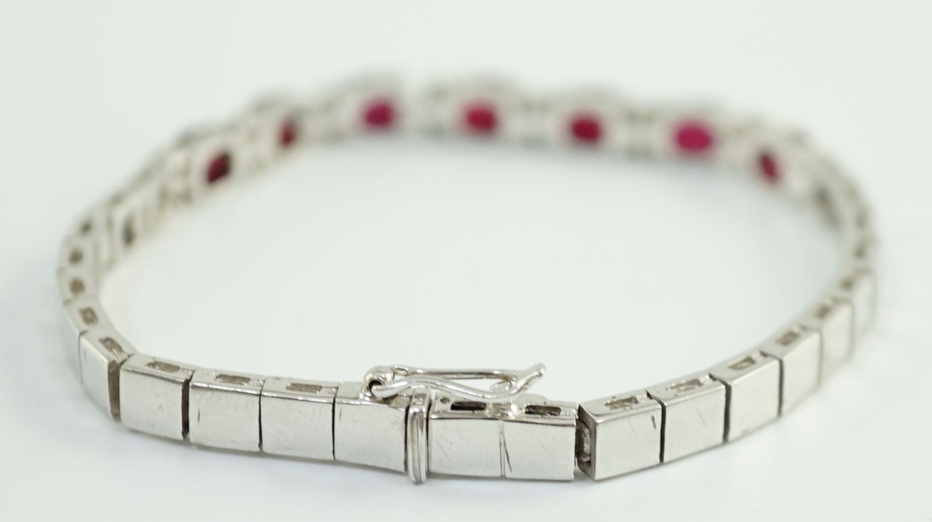 A 750 white gold and seven stone oval cut ruby set line bracelet, set with sixteen stone diamond spacers